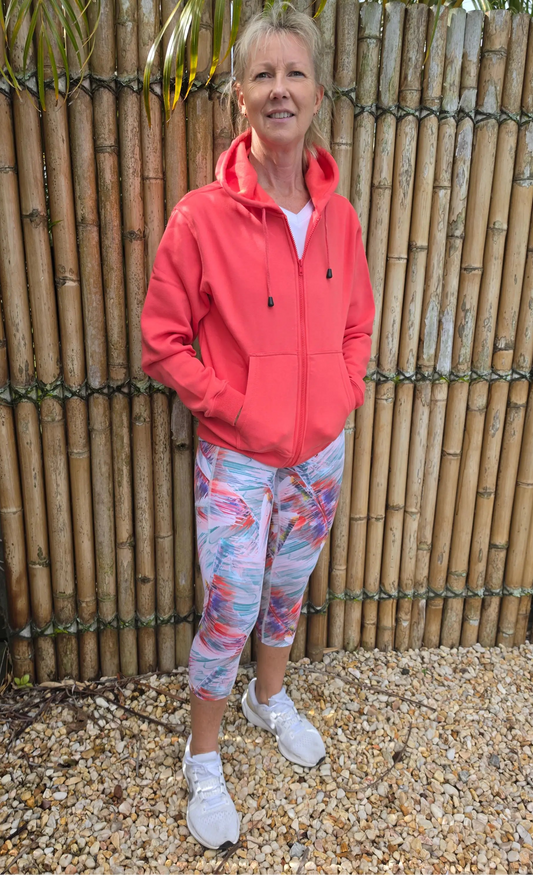 Coral-colored zip-up hoodie with hood and pockets worn over white top paired with moisture wicking active leggings